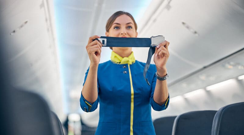 Close up portrait of young cheerful flight attendant in blue business uniform demonstrating how fostering passenger seatbelt